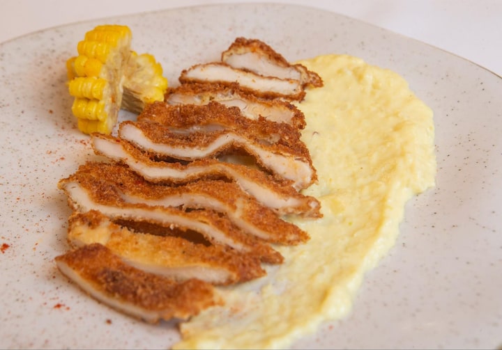 Filé de frango à milanesa com creme de milho, servido em um prato raso de cor bege, com bordas douradas. O prato está servido em cima de uma mesa branca.