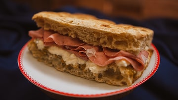 Sanduíche de pastrami no pão de forma integral sobre prato de louça branca com listra vermelha na borda. Foto: John Alves