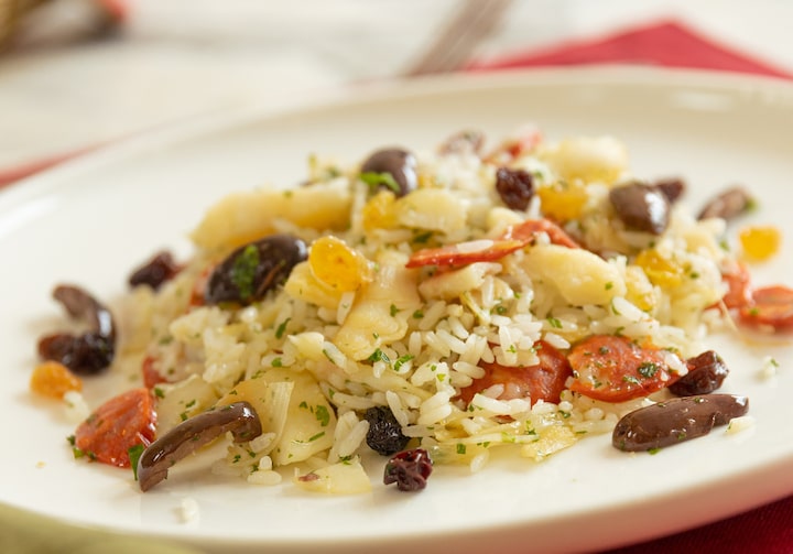 Em um prato branco, lascas de bacalhau, azeitonas pretas e uvas-passas estão em cima de uma porção de arroz branco. O prato está sobre um jogo-americano vermelho.