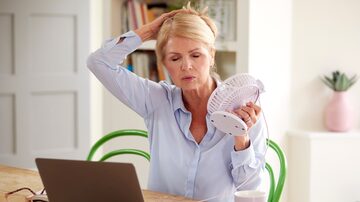 Menopausal Mature Woman Having Hot Flush At Home Cooling Herself With Fan Connected To Laptop. Foto: Monkey Business/Adobe Stock