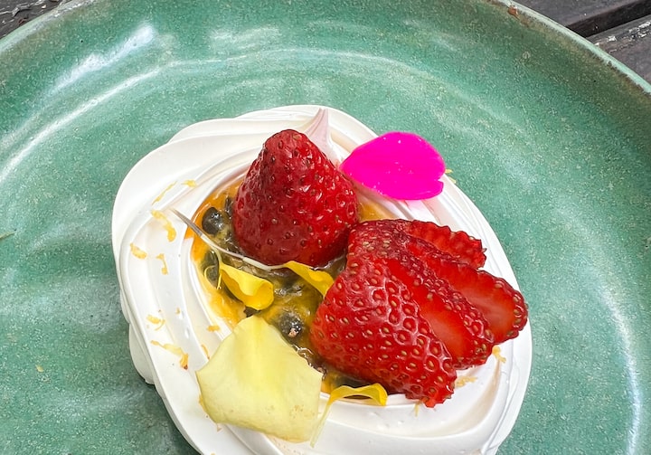 Pavlova com base branca, creme de maracujá e morangos em cima - servida em um prato raso, com bordas altas, na cor verde. O prato está servido em uma mesa de madeira.