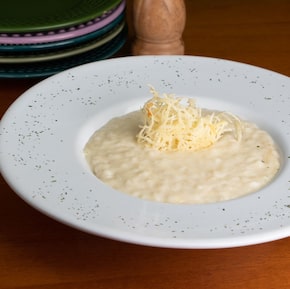 Em um prato fundo de louça branco, está o risoto cremoso de quatro queijos com queijo ralado ao centro, por cima. Foto: Frutaria Pitaya/Divulgação