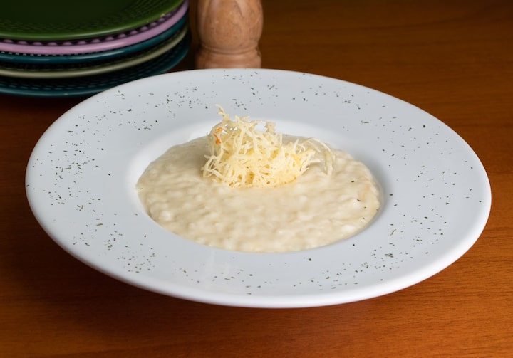 Em um prato fundo de louça branco, está o risoto cremoso de quatro queijos com queijo ralado ao centro, por cima.