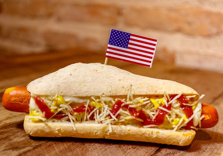 Pão de queijo recheado com salsicha e coberto por batata palha, ketchup e mostarda, e uma bandeirinha dos Estados Unidos fincada no topo