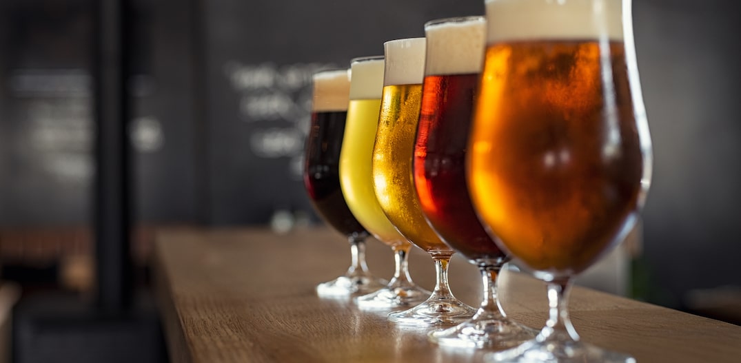 Sobre balcão de madeira e em foco, 5 taças de cerveja de diferentes tipos e cores dispostas enfileiradas. Foto: Rido/Adobe Stock