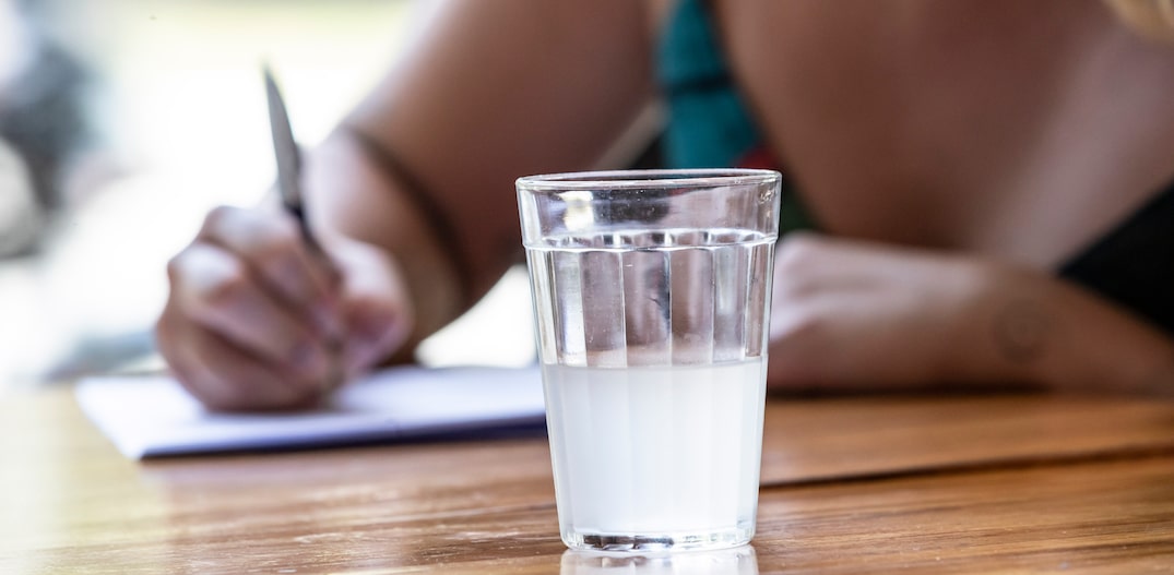 Mulher aparece parcialmente ao fundo da imagem fazendo anotações em um papel branco. A sua frente, um copo americano com água de coco. Foto: TABA BENEDICTO