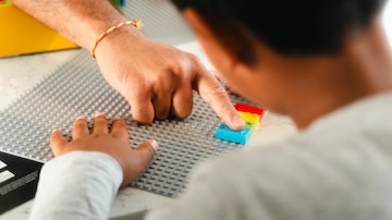 Mão esticada toca com um dedo a parte de cima de uma peça de Lego Braille Bricks, onde ficam os pinos que representam uma letra ou número em Braille; em primeiro plano, uma criança de costas para a câmera, virada para o Lego. Foto: Divulgação/Lego