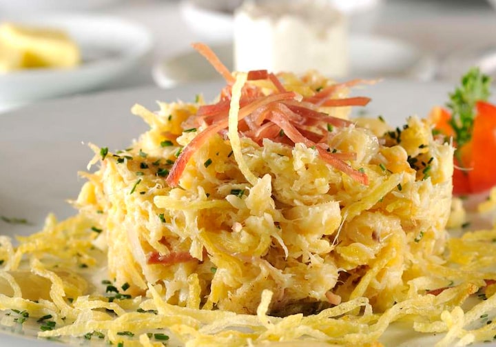 Em um prato branco está servido o bacalhau com batata palha e presunto de Parma. Atrás, há outros pratinhos - um com queijo amarelo e outro com queijo branco. Fundo desfocado.
