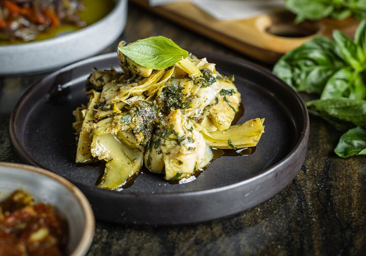 Em foco, uma porção da alcachofra marinada, servida com molho de ervas , alho, e raspas de limão siciliano sobre um prato de cerâmica preta de bordas altas.