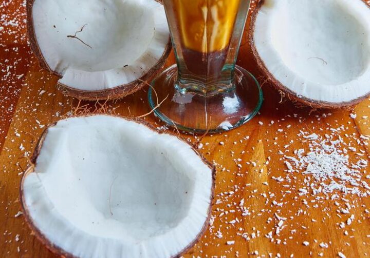Uma taça de milkshake de coco com calda de caramelo, disposta em uma mesa de madeira com cocos abertos e raspas de coco.