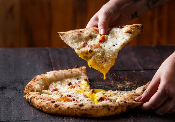 Em uma mesa de madeira está uma pizza artesanal de carbonara.