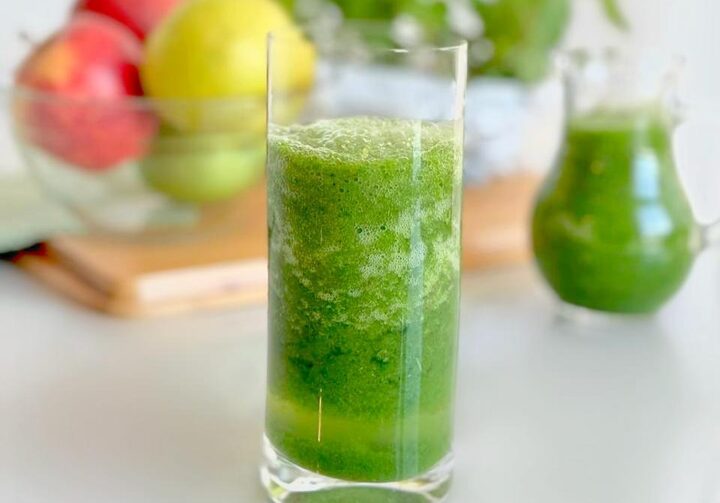 Em uma mesa branca está um copo alto e transparente com suco verde. Ao fundo, frutas.