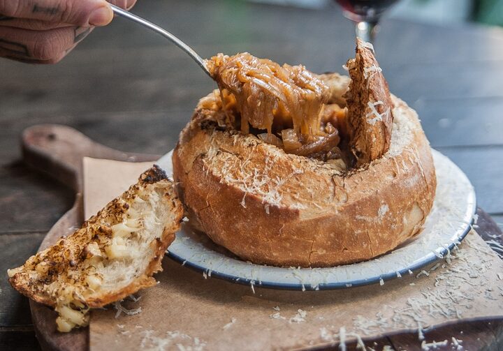 Imagem mostra uma taça de vinho tinto e ao lado um prato de sopa de cebola servida dentro de um pão italiano redondo com uma pessoa se servindo