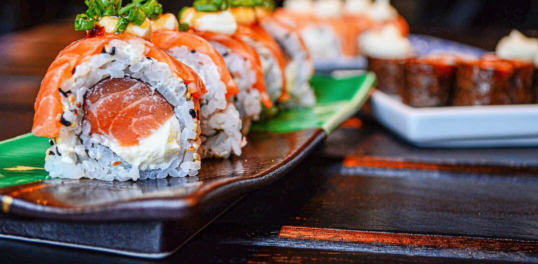 Sushi de salmão enrolado em arroz e alga, com mais uma peça de salmão em cima, servidos em uma tigela vermelha. Foto: Unsplash