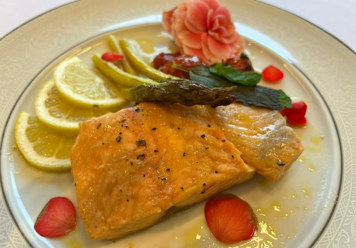 Em um prato branco, uma posta de salmão servida com aspargos, fatias de limão siciliano e pétalas de flor comestível na cor rosa.