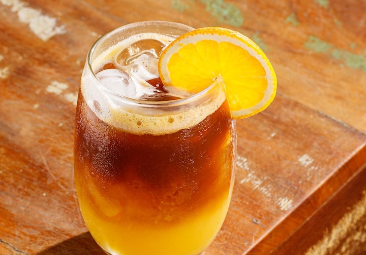 Café com laranja servido em um copo Aruba, bojudo, decorado com muito gelo e uma rodela de laranja na borda. O copo está em uma mesa de madeira, com detalhes em branco e verde.