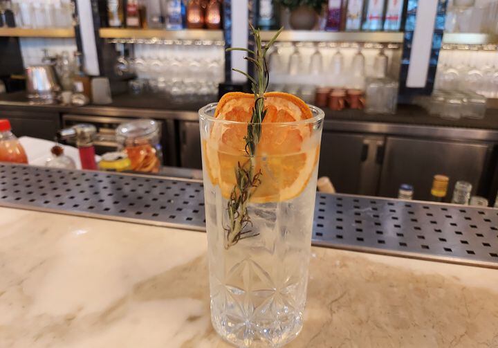 Sobre uma bancada de pedra branca, está um copo longo de cristal transparente contendo o drinque Monte Carlo Fizz, feito com água tônica. Enfeitando, um ramo de alecrim e uma laranja desidratada.